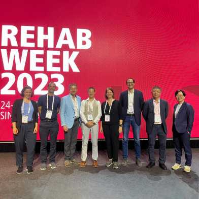 Eight scientists posing in front of a huge display at REHAB Week 2023