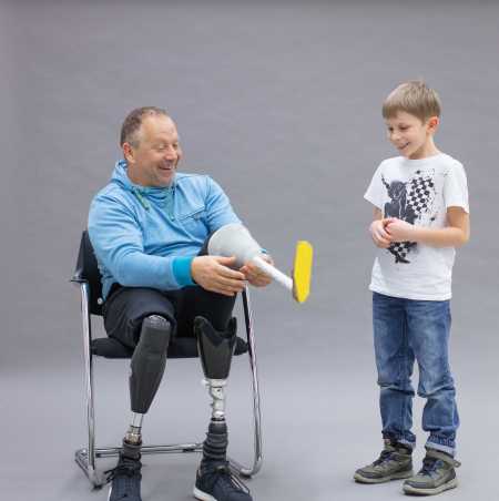 Enlarged view: Rüdiger Böhm with two prosthetic legs is laughing and trying a handmade prosthesis made of paper, which he receives from a boy.
