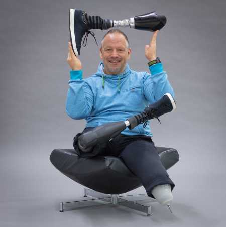Enlarged view: Rüdiger Böhm with two prosthetic legs sits on armchair and holds prosthesis in victory pose over head