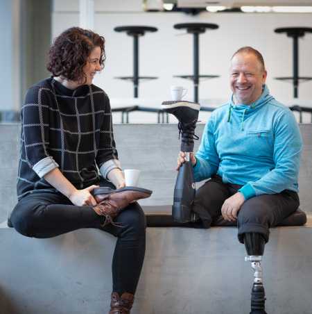 Enlarged view: Rüdiger Böhm with two prosthetic legs sits opposite a woman. Both balance a coffee cup also on their shoe